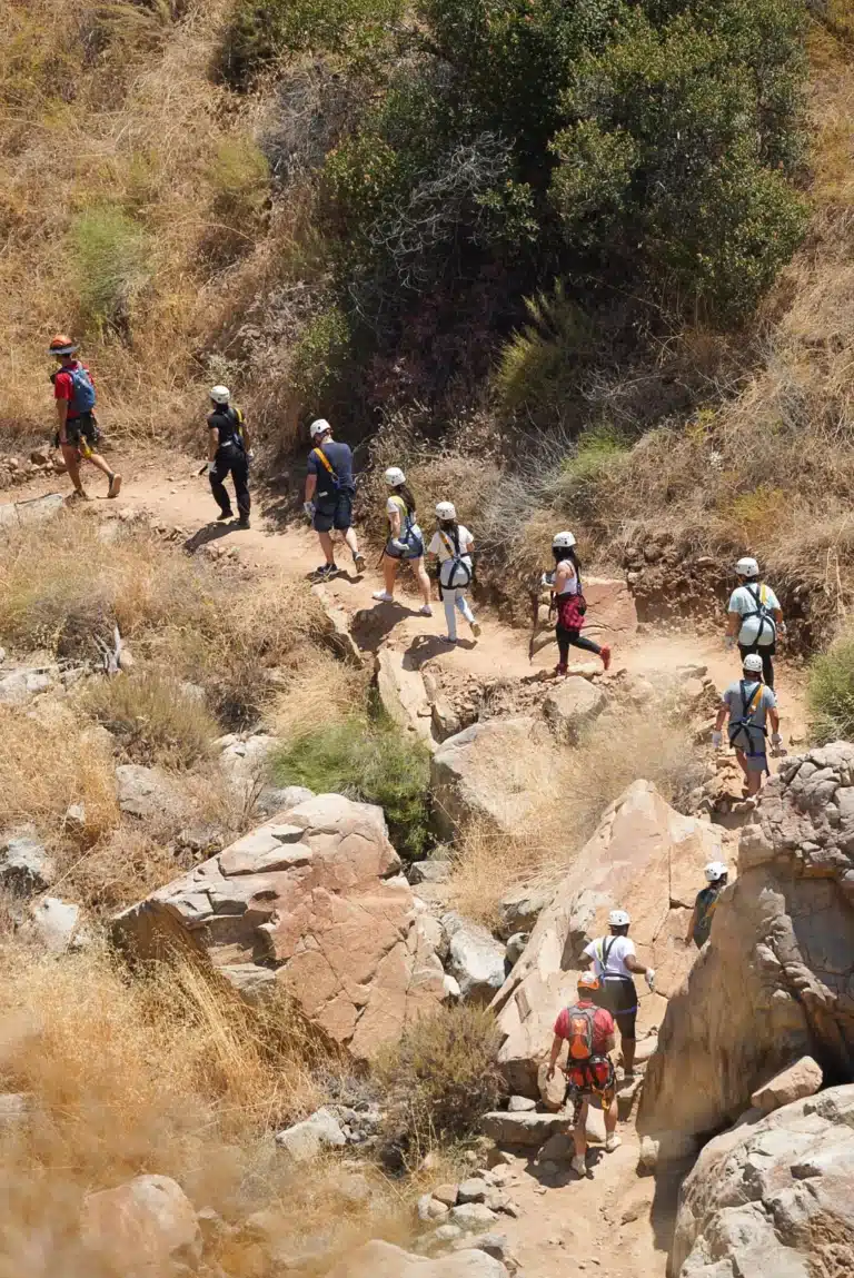 A group of people wearing helmets and backpacks hike along a rocky, dirt trail surrounded by dry grass and shrubs.