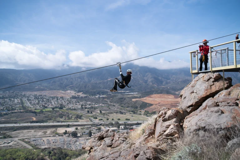 A person rides a zipline over a rocky hill, with another person standing on a platform; a town and mountains are visible in the background.