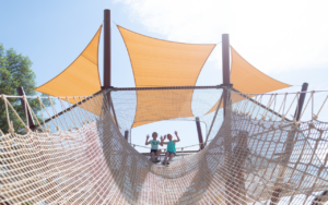 Two children stand on a rope bridge under orange shade sails, waving and smiling like heroes in a playground sandbox on a sunny day.