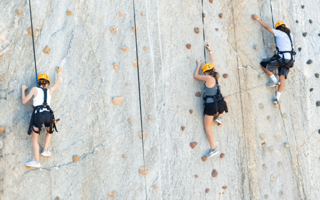 Three people wearing helmets and harnesses climb a vertical outdoor rock wall with safety ropes attached, showing true 2025 Hero spirit as they tackle the challenge like a real-life adventure sandbox.