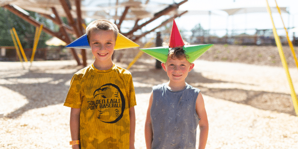 Two children standing outdoors, each wearing colorful cone-shaped hats and smiling at the camera on a sunny day—perfect for capturing Summer Birthday Party Sale memories.